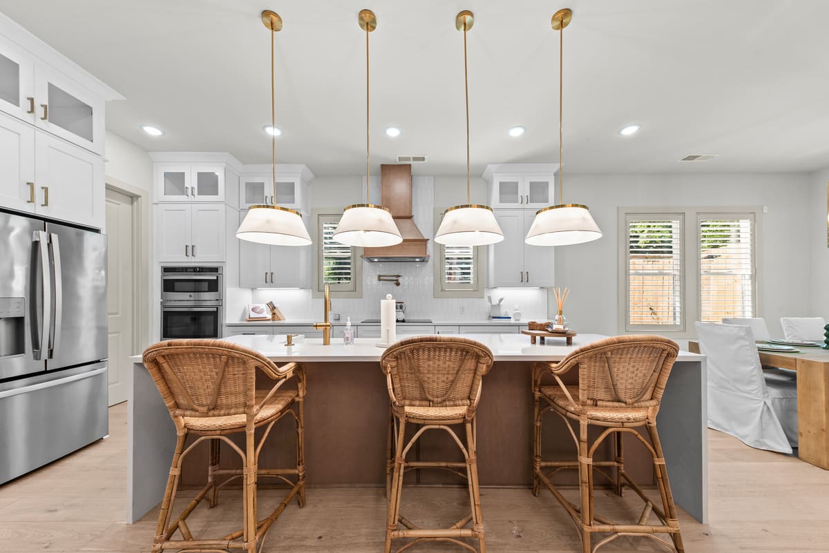 Bright white kitchen with large island, three rattan counter stools, and four brass pendant lights above a quartz countertop