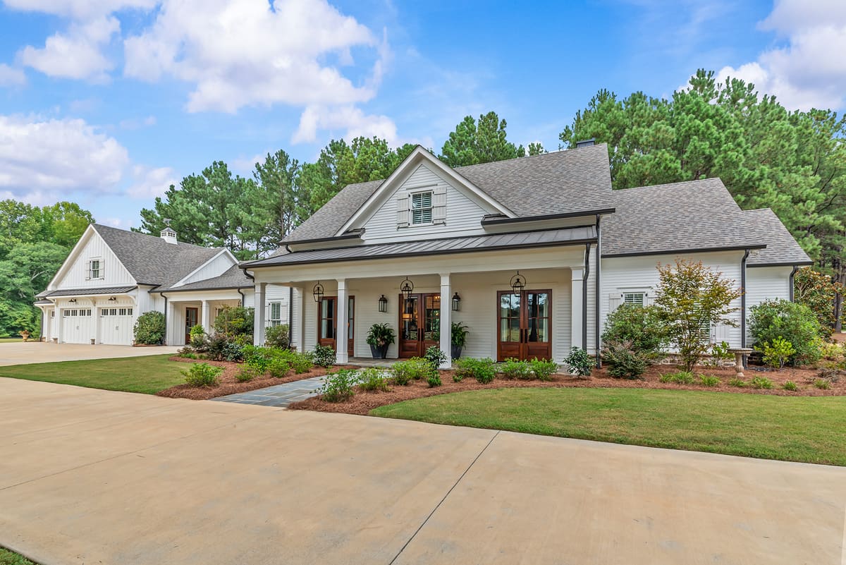 Front elevation of a white modern farmhouse with covered porch, wood doors, and attached garage surrounded by mature pines